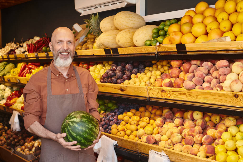 vreugdevolle goed uitziende verkoper met baard poseren met verse watermeloen in handen en glimlachen op de camera - Foto, afbeelding