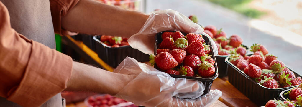 bijgesneden beeld van volwassen verkoper houden verpakking van verse aardbeien in handen door het raam in de winkel, banner - Foto, afbeelding
