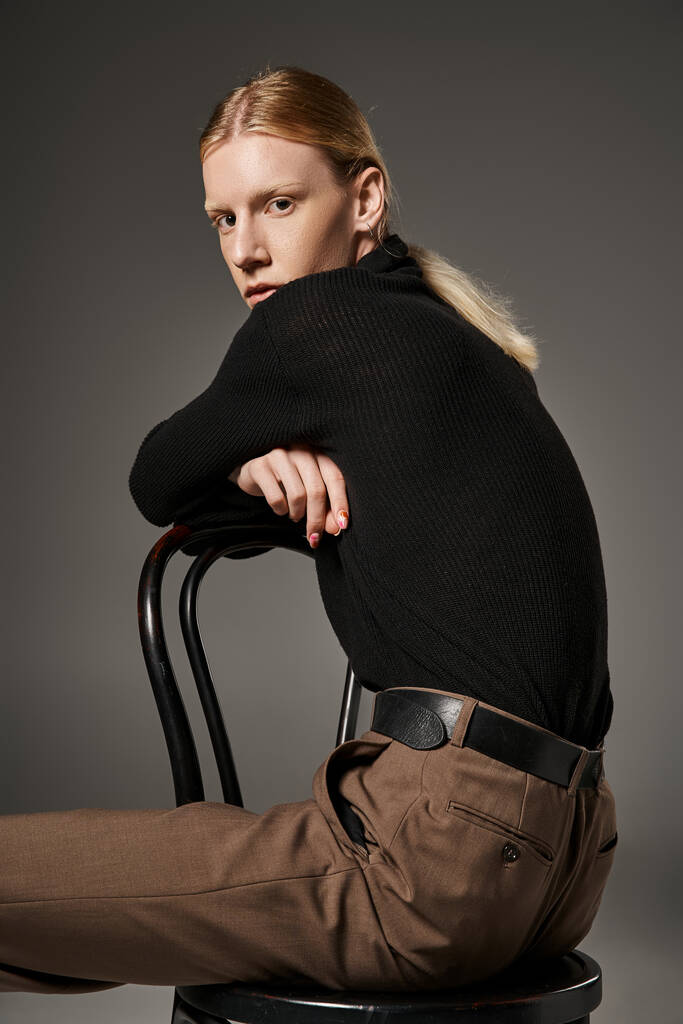 verticaal shot van stijlvol non-binair model in trendy outfit zittend op stoel en kijkend naar camera - Foto, afbeelding