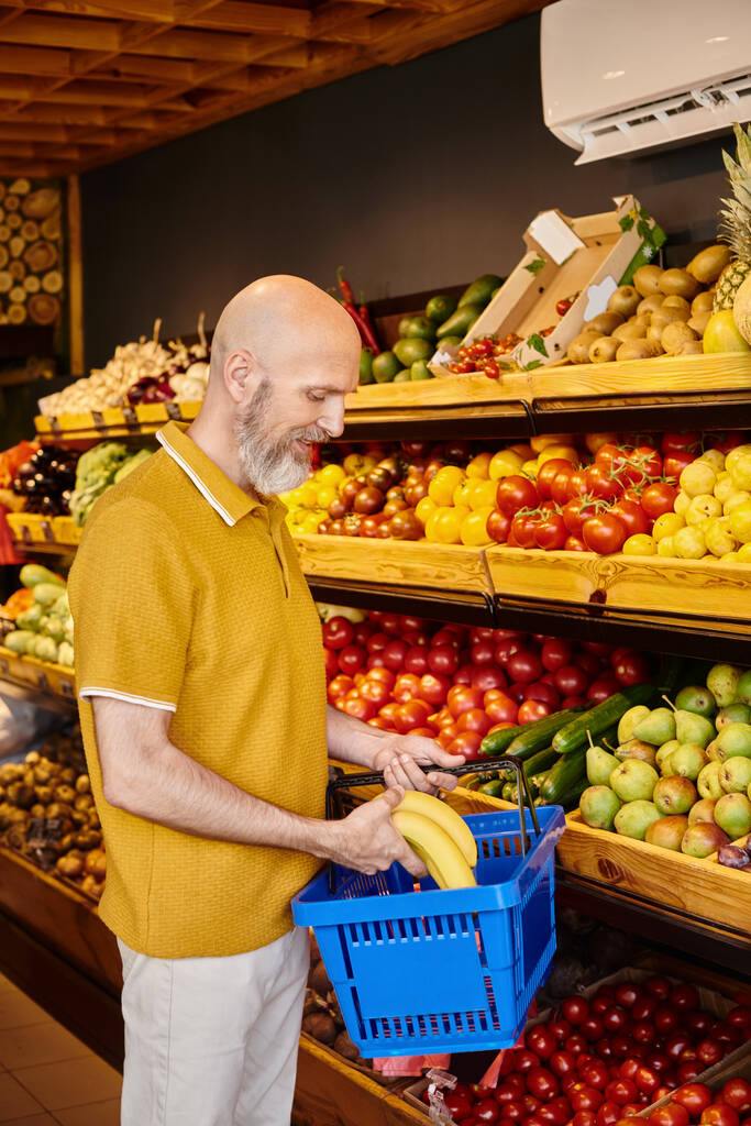 cheerful mature customer in casual attire putting fresh vibrant bananas into shopping basket - Photo, Image