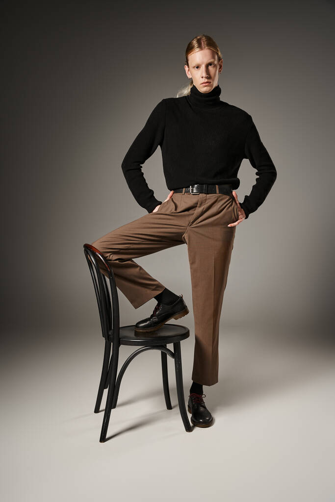 вертикальный снимок небинарного человека в модном наряде с ногой на стуле и руками в карманах - Фото, изображение