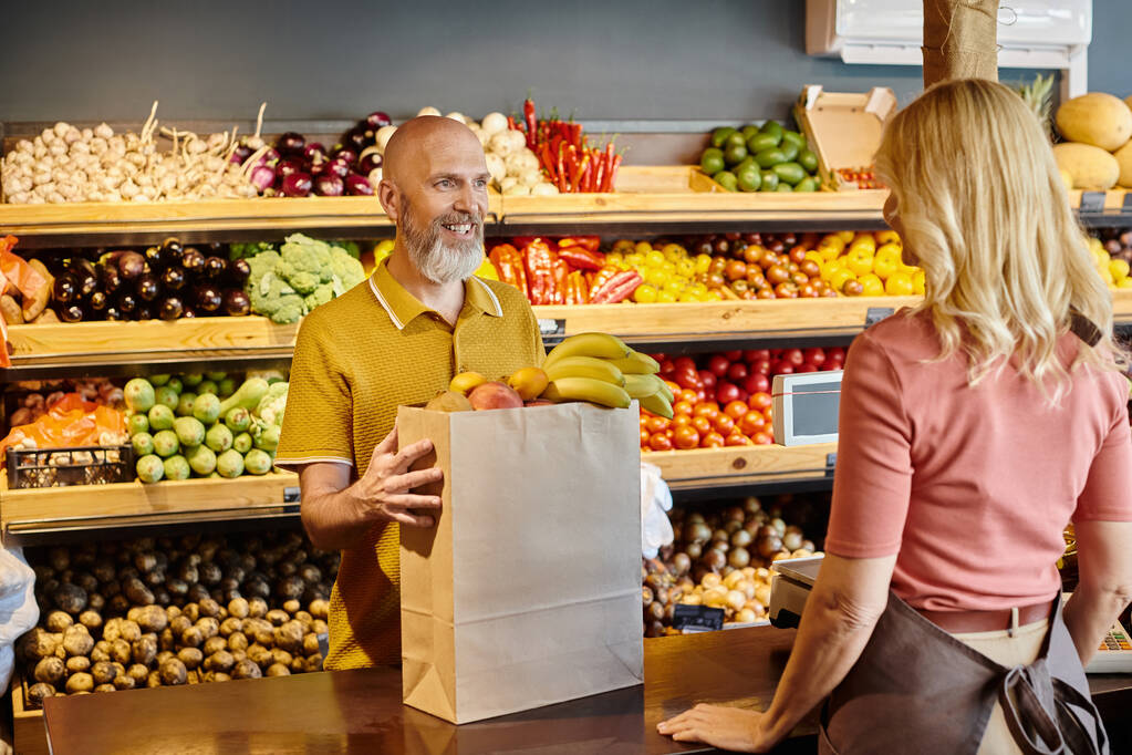 centrarse en el cliente maduro comprar frutas frescas y sonreír al vendedor femenino borroso en la tienda de comestibles - Foto, imagen