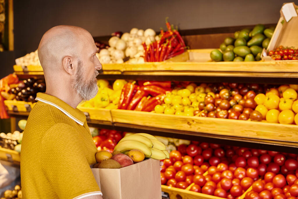 concentrated mature gray bearded man holding paper bag full of fresh fruits at grocery store - Photo, Image