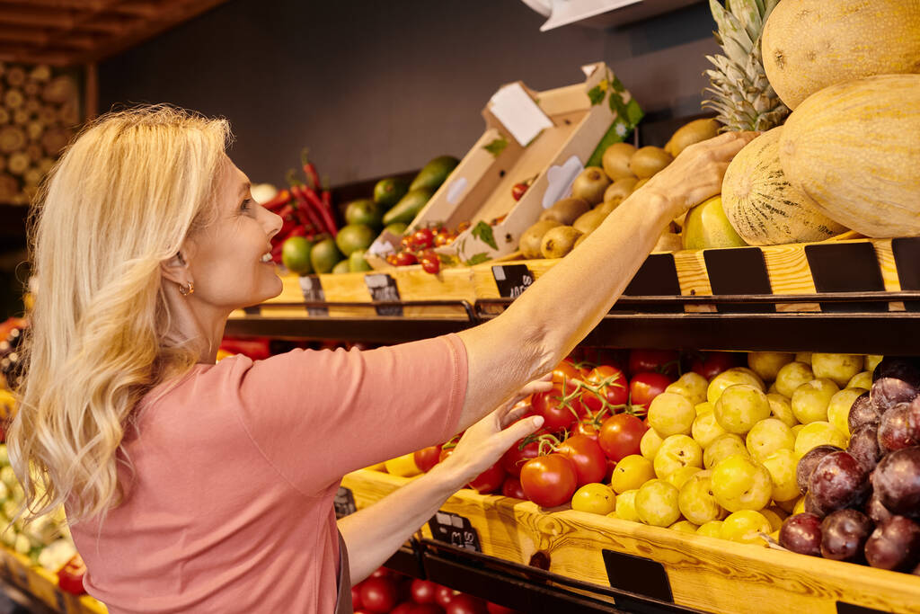 dojrzały wesoły sprzedawca uśmiechnięty podczas pracy obok stoiska spożywczego ze świeżymi owocami i warzywami - Zdjęcie, obraz