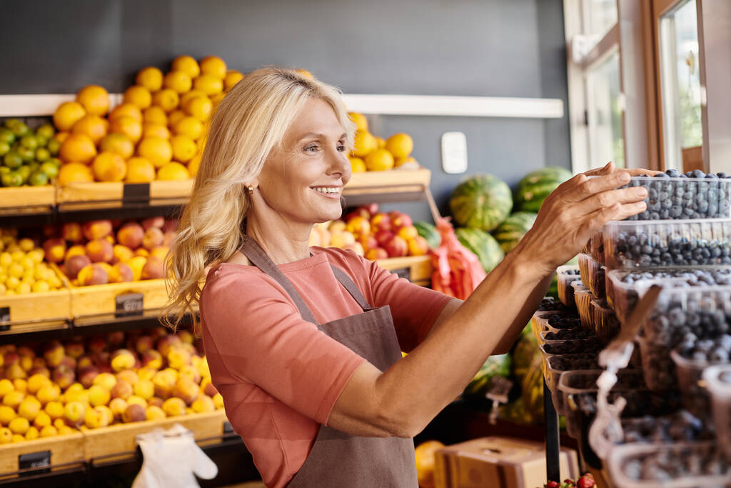 fröhlicher älterer Verkäufer überprüft Packungen mit Brombeeren mit Obststand im Hintergrund im Lebensmittelladen - Foto, Bild