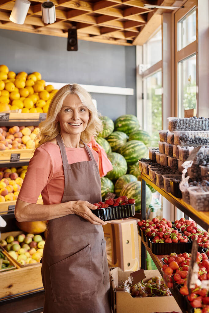 alegre madura vendedora sosteniendo deliciosas fresas frescas y sonriendo felizmente a la cámara - Foto, imagen