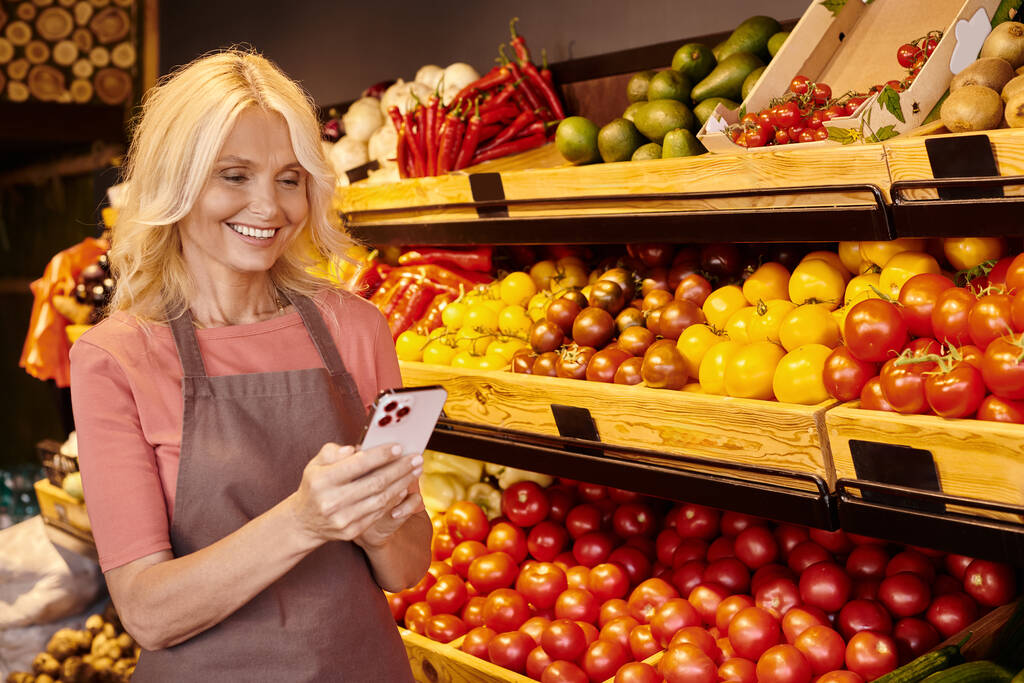 cheerful mature saleswoman smiling happily and looking at her mobile phone at grocery store - Photo, Image