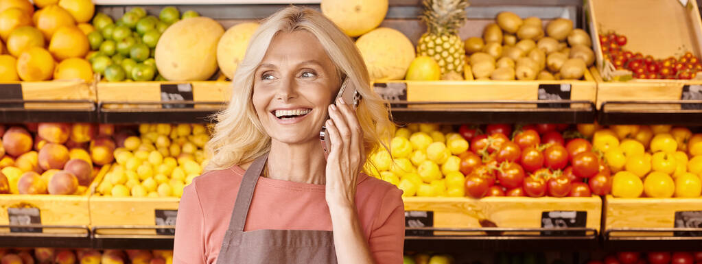 happy female seller smiling and talking by phone with fruits and vegetables on backdrop, banner - Photo, Image
