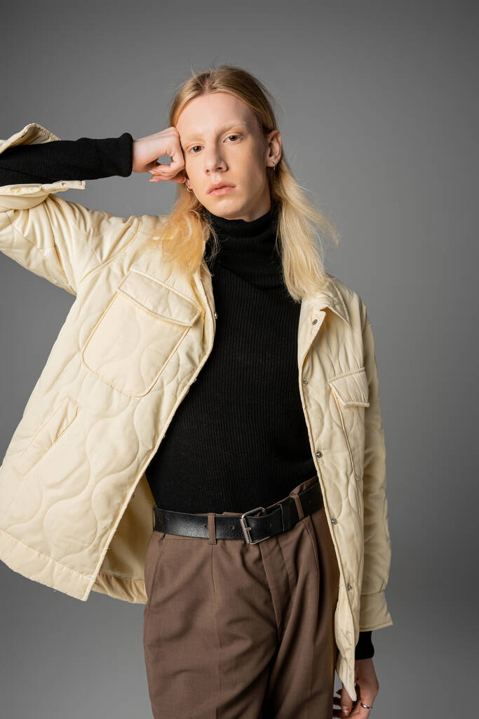 mladý pohledný nebinární model ve stylovém teplém oblečení pózující s rukou zvednutou k hlavě, móda - Fotografie, Obrázek