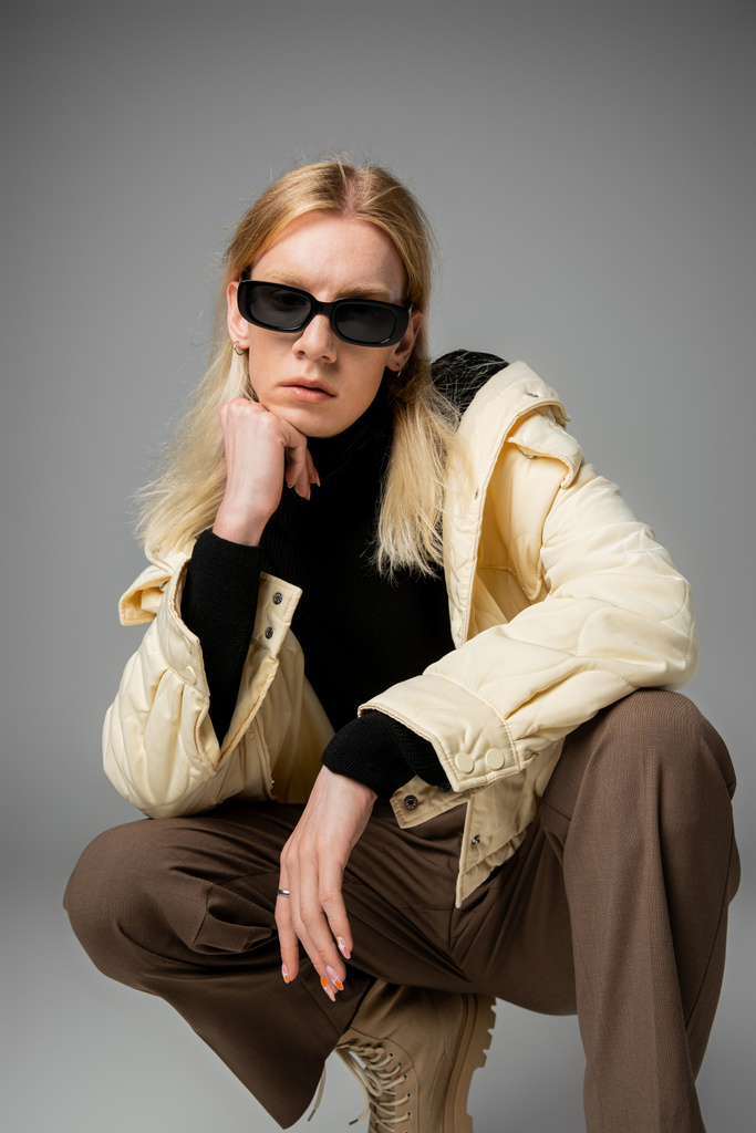 atractiva persona andrógina en traje elegante con gafas de sol en cuclillas con puño debajo de la barbilla - Foto, Imagen