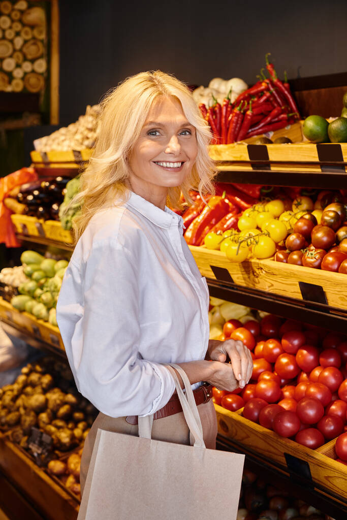 maduro cliente alegre con bolsa de papel mirando felizmente a la cámara al lado de frutas y verduras frescas - Foto, Imagen
