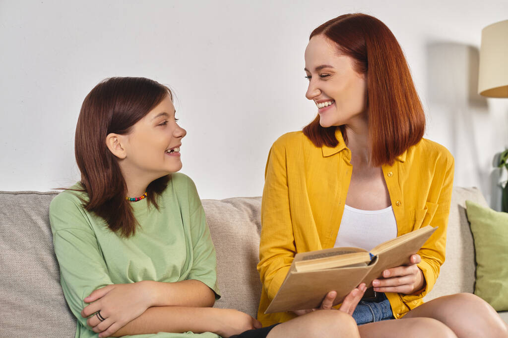 улыбающаяся женщина читает книгу о половом воспитании дочери-подростка на уютном диване в гостиной - Фото, изображение