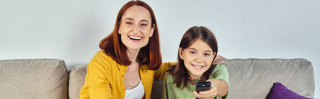 śmiejąca się kobieta z nastoletnią córką oglądającą film na przytulnej kanapie w nowoczesnym salonie, baner - Zdjęcie, obraz