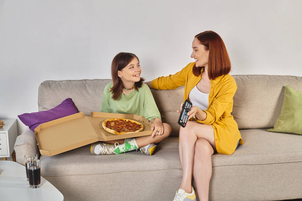 happy mother and teen daughter watching tv on couch near pizza and glasses with soda in living room - Photo, Image