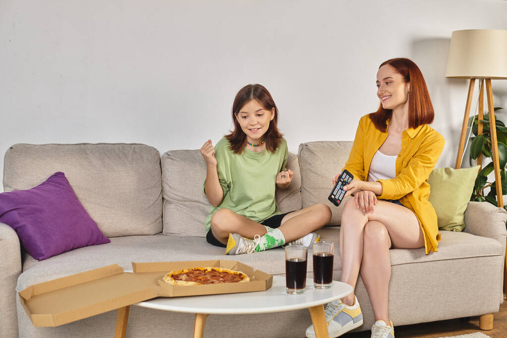 feliz madre e hija adolescente viendo la televisión en el sofá cerca de pizza y vasos con soda en la sala de estar - Foto, imagen