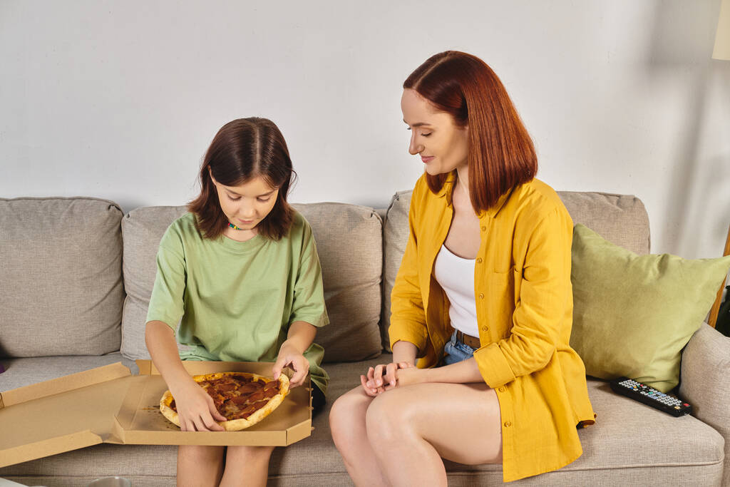 дівчина сидить на дивані зі смачною піцою біля усміхненої матері на дивані у вітальні - Фото, зображення