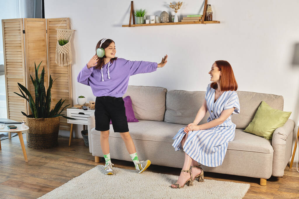 fröhliches Teenager-Mädchen mit Kopfhörern, das Musik hört und neben lächelnder Mutter im Wohnzimmer tanzt - Foto, Bild