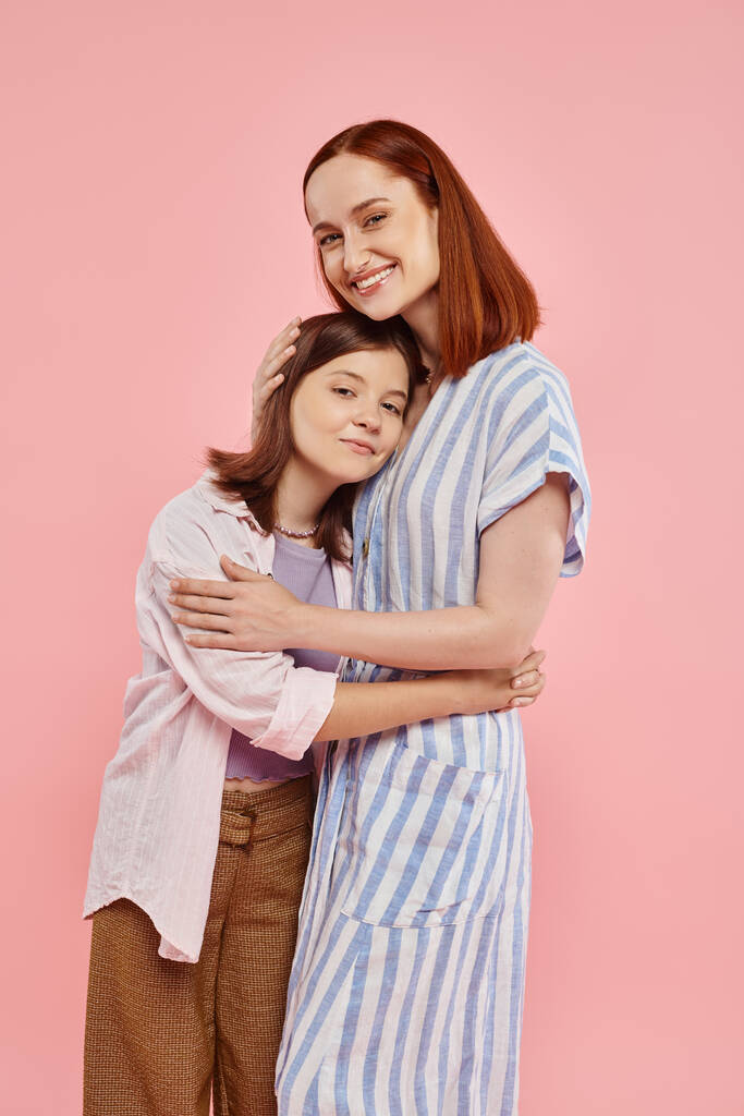 mujer feliz con hija adolescente en elegante atuendo casual abrazando y mirando a la cámara en rosa - Foto, imagen