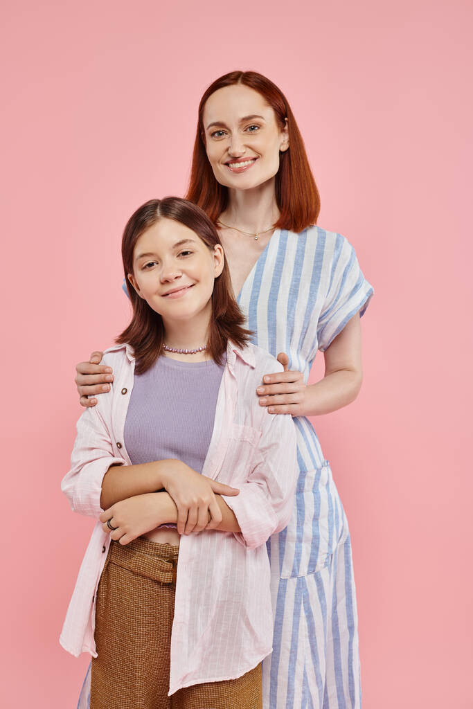 fröhliche und stilvolle Frau umarmt Schultern ihrer Teenager-Tochter und schaut in die Kamera auf rosa - Foto, Bild