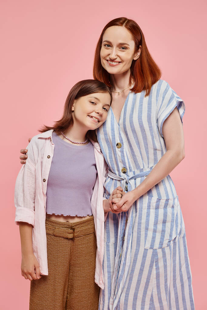 mujer alegre y elegante con hija adolescente cogida de la mano y sonriendo a la cámara en el telón de fondo rosa - Foto, Imagen