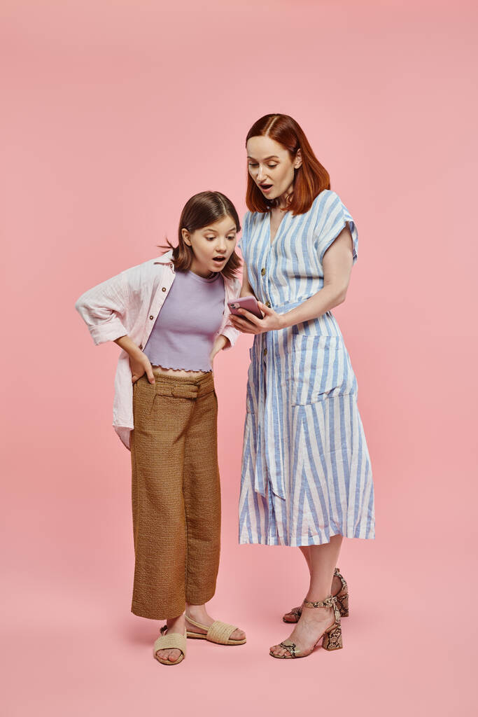 hämmästynyt äiti ja teini tytär katselee matkapuhelinta seisoessaan vaaleanpunaisella taustalla - Valokuva, kuva
