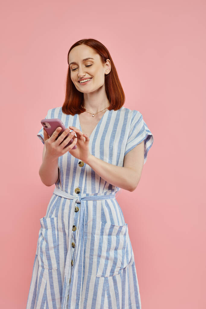 весела і стильна жінка в смугастій сукні з використанням мобільного телефону на рожевому тлі, сучасний спосіб життя - Фото, зображення