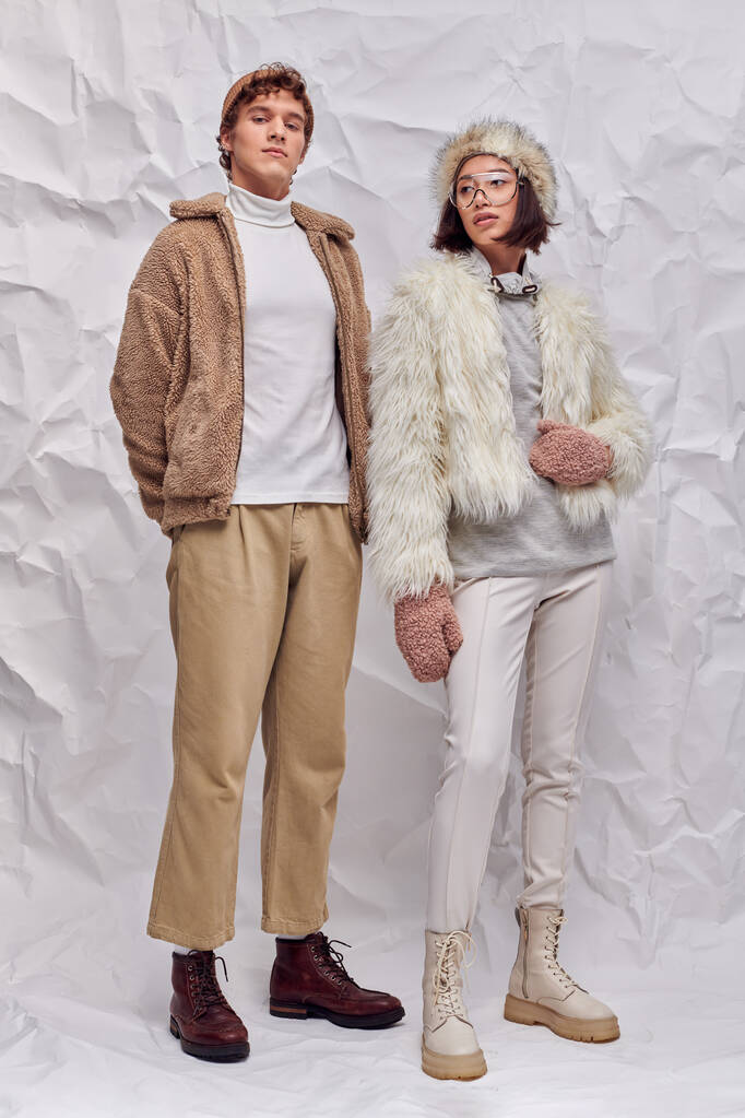 trendy man op zoek naar camera in de buurt van aziatische vrouw in warme jas en wanten op witte textuur achtergrond - Foto, afbeelding