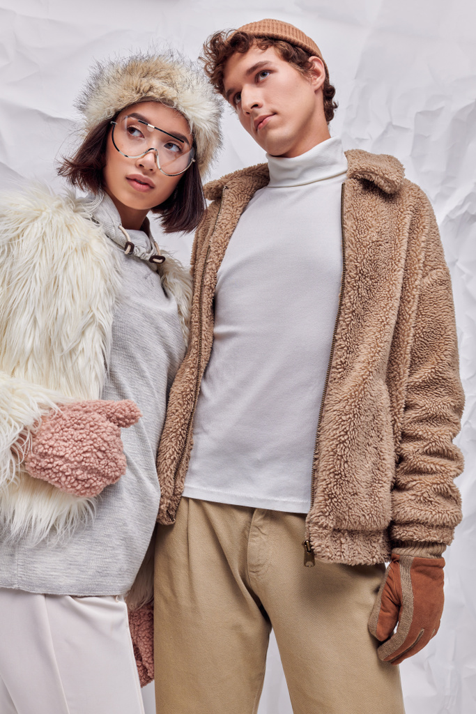 зимняя мода, межрасовая пара в теплой верхней одежде глядя в сторону на белом текстурированном фоне - Фото, изображение