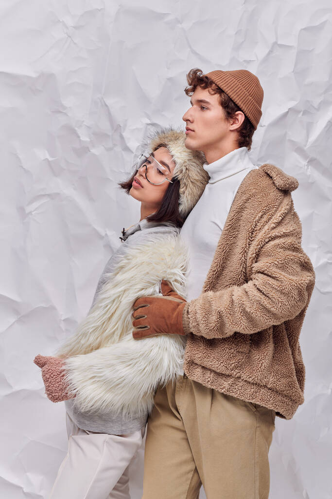 trendy man in warm clothes hugging young asian model and looking away on white crumpled backdrop - Photo, Image