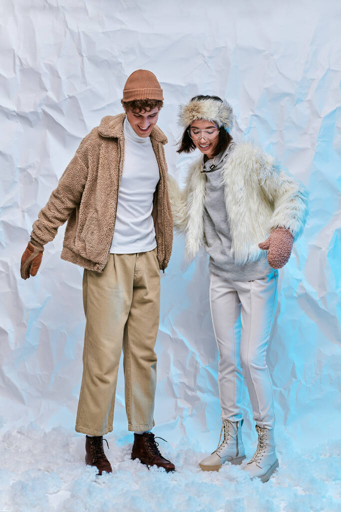 joyful interracial couple in trendy winter clothes standing on snow on white textured backdrop - Photo, Image