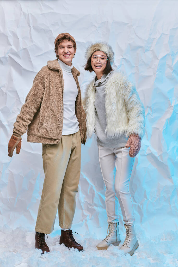 excited interracial couple in warm stylish clothes standing on snow in studio, winter fashion - Photo, Image