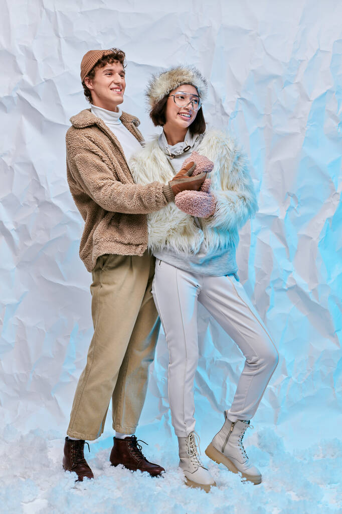 Unbekümmertes gemischtrassiges Paar in stylischer Winterkleidung, das auf Schnee vor weiß strukturiertem Hintergrund wegschaut - Foto, Bild