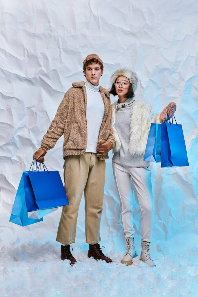 modna para międzyrasowa z niebieskimi torbami na zakupy na białym śniegu w studio, zakupy zimowe - Zdjęcie, obraz