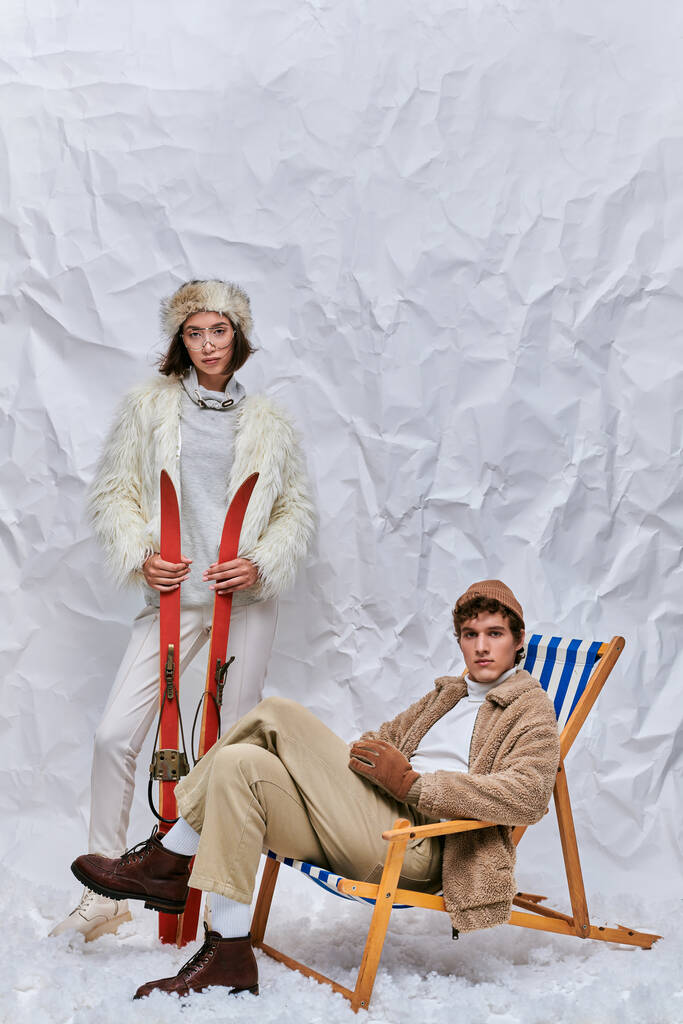 invierno moda y ocio, asiático mujer con esquís cerca de moda hombre en cubierta silla en snowy studio - Foto, Imagen