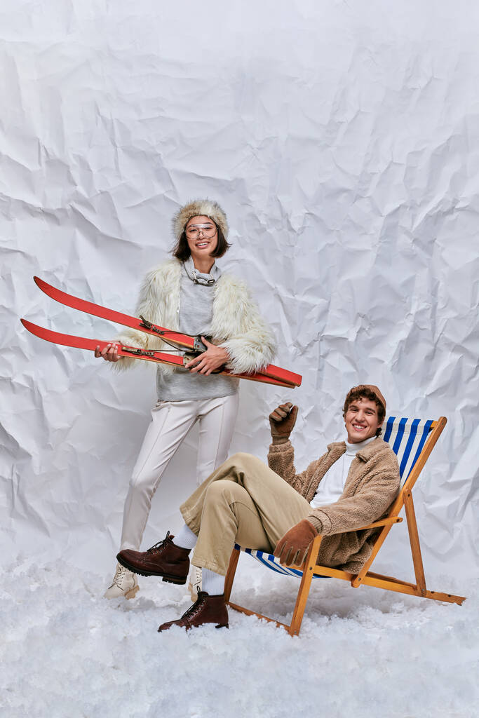 cheerful asian woman with skis near trendy man in deck chair on snow in studio, winter leisure - Photo, Image