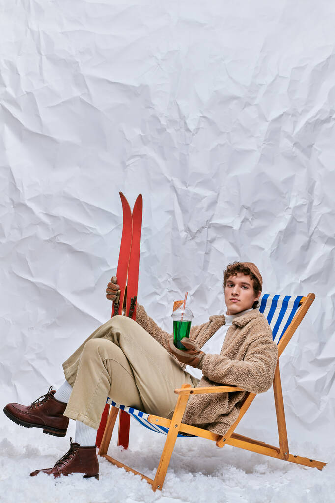 modny mężczyzna w ciepłym stroju siedzi na leżaku z gorącym koktajlem i napojem w zaśnieżonym studio - Zdjęcie, obraz