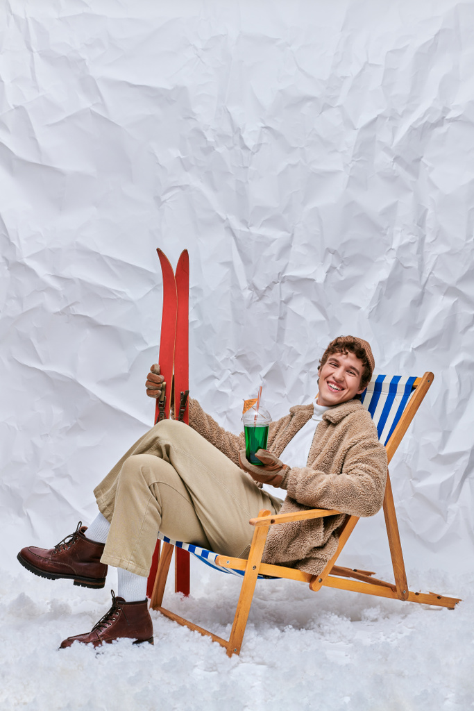homem alegre no traje do inverno que senta-se na cadeira da plataforma com bebida do apres-esqui e esquis no estúdio nevado - Foto, Imagem