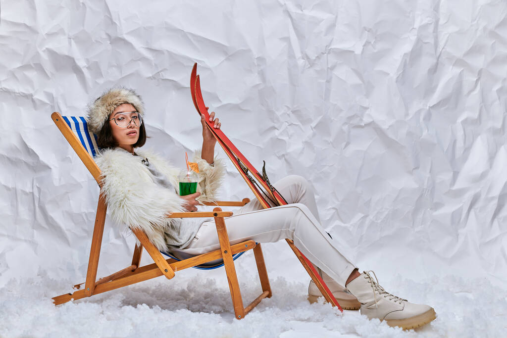 aziatische vrouw in de winter dragen zitten in dek stoel met ski 's en hete teddy cocktail op sneeuw in studio - Foto, afbeelding