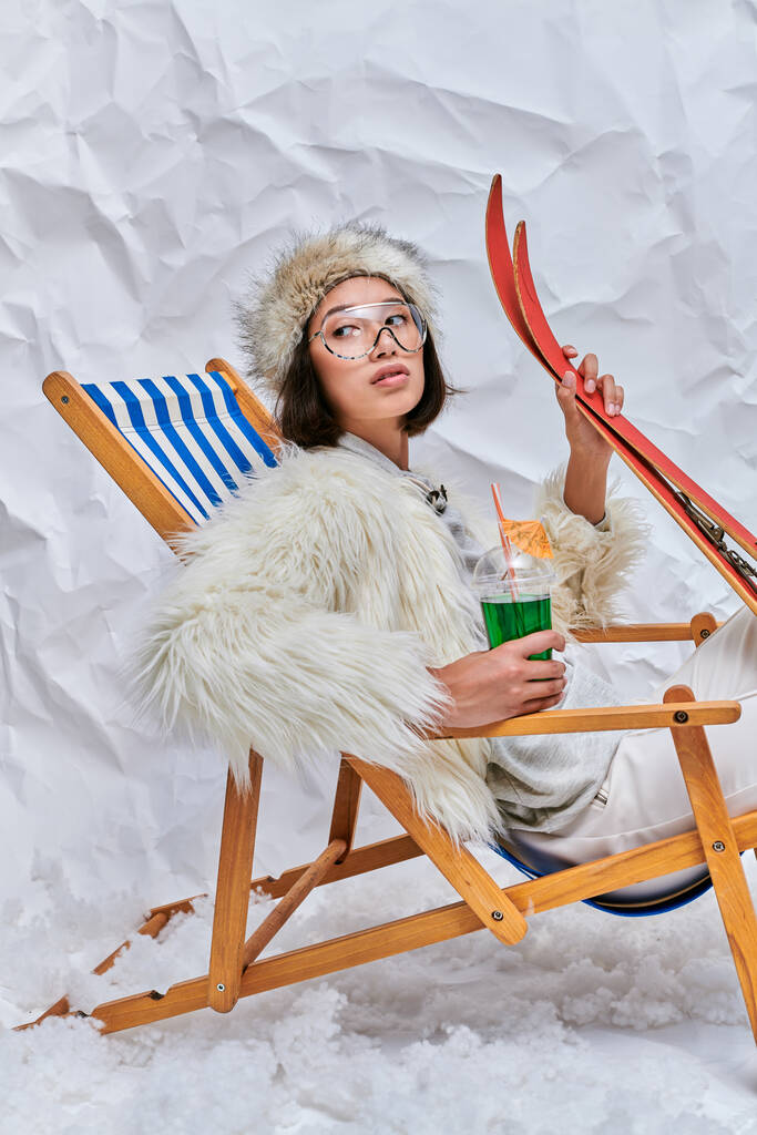 mujer asiática en gafas y ropa de invierno relajante en tumbona con cóctel y esquís en estudio - Foto, imagen
