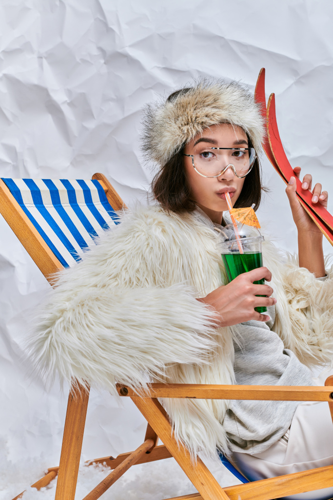 白質の背景にデッキチェアでホットテディカクテルを飲むスキーを持つアジアの女性 - 写真・画像