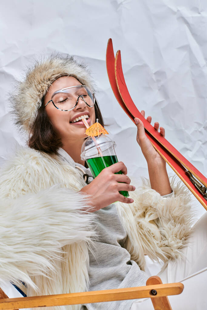 joyful asian model with skis drinking apres-ski cocktail in deck chair on white textured backdrop - Photo, Image