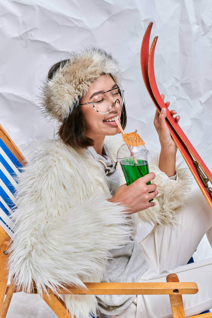 щаслива азіатка з лижами п'є гарячий плюшевий коктейль із закритими очима на білому текстурованому тлі - Фото, зображення