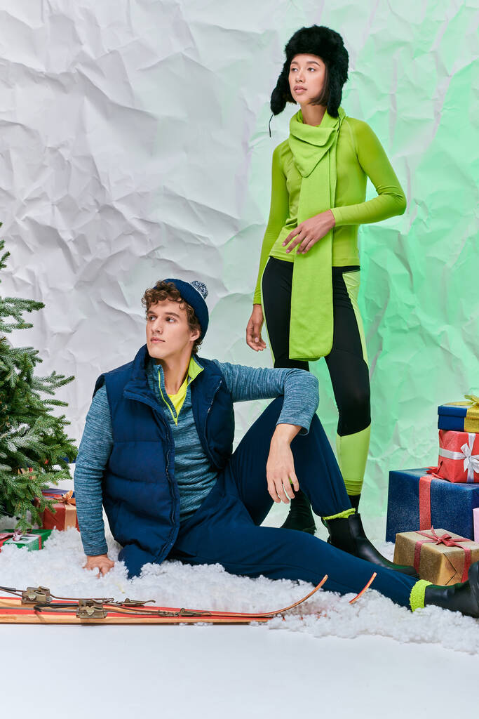 διαφυλετικό ζευγάρι το χειμώνα φορούν κοιτάζοντας μακριά δίπλα σε δώρα και χριστουγεννιάτικο δέντρο σε χιονισμένο στούντιο - Φωτογραφία, εικόνα