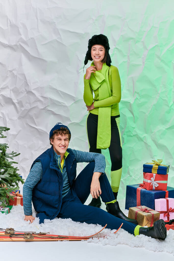 międzyrasowa para w stroju zimowym uśmiechnięta w pobliżu choinki, narty i prezenty w zaśnieżonym studio - Zdjęcie, obraz