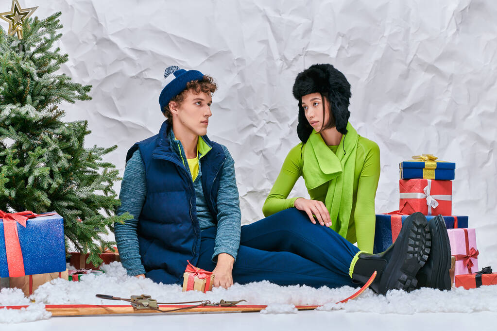 modieuze interraciale paar zitten op sneeuw in de studio in de buurt van ski 's, cadeautjes en kerstboom - Foto, afbeelding