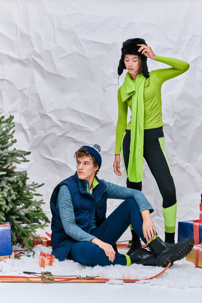 homme à la mode assis sur la neige en studio près de la femme asiatique, boîtes-cadeaux et arbre de Noël, mode d'hiver - Photo, image