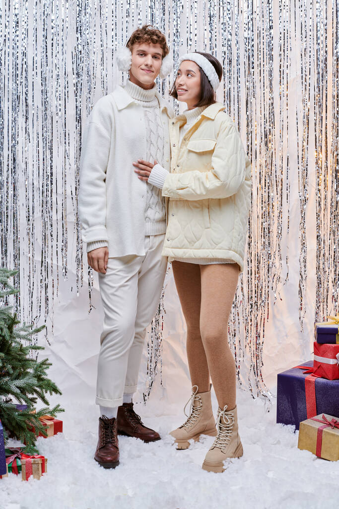 glücklich asiatische Frau Blick auf stilvollen Mann in der Nähe von Geschenken und Weihnachtsbaum auf glänzendem Lametta Hintergrund - Foto, Bild