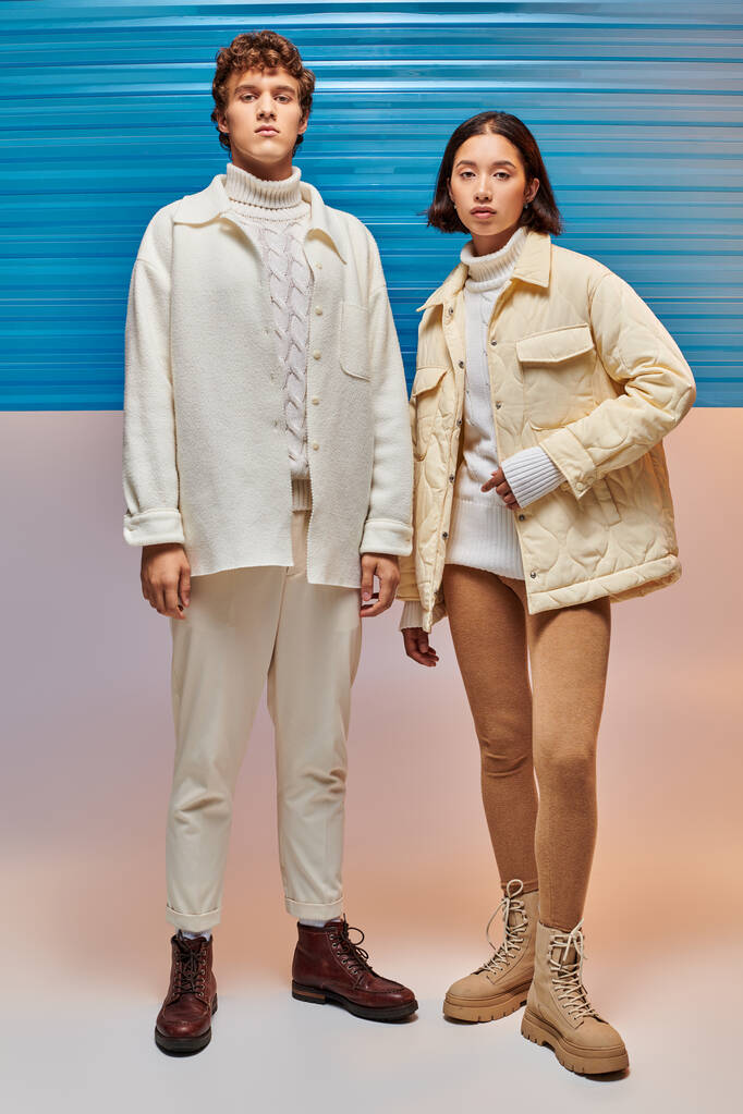 interraciale paar in warme jassen en lederen laarzen op zoek naar camera in de buurt van blauw plastic paneel - Foto, afbeelding