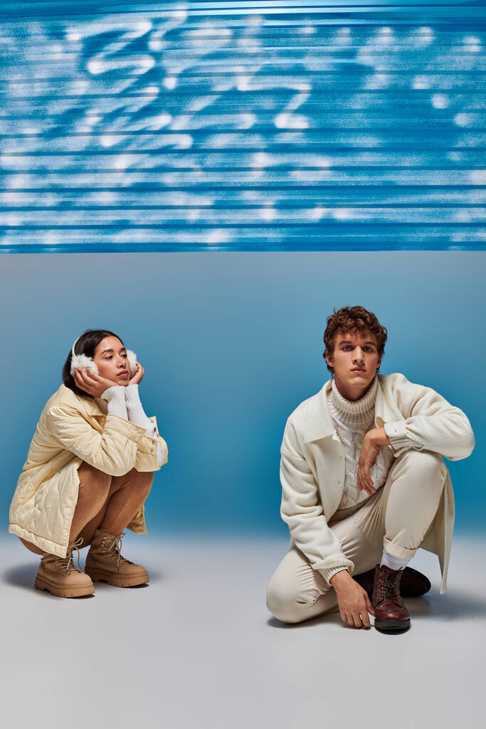 pareja interracial vestida a la moda posando en haunches cerca de la lámina de plástico azul en el estudio - Foto, imagen