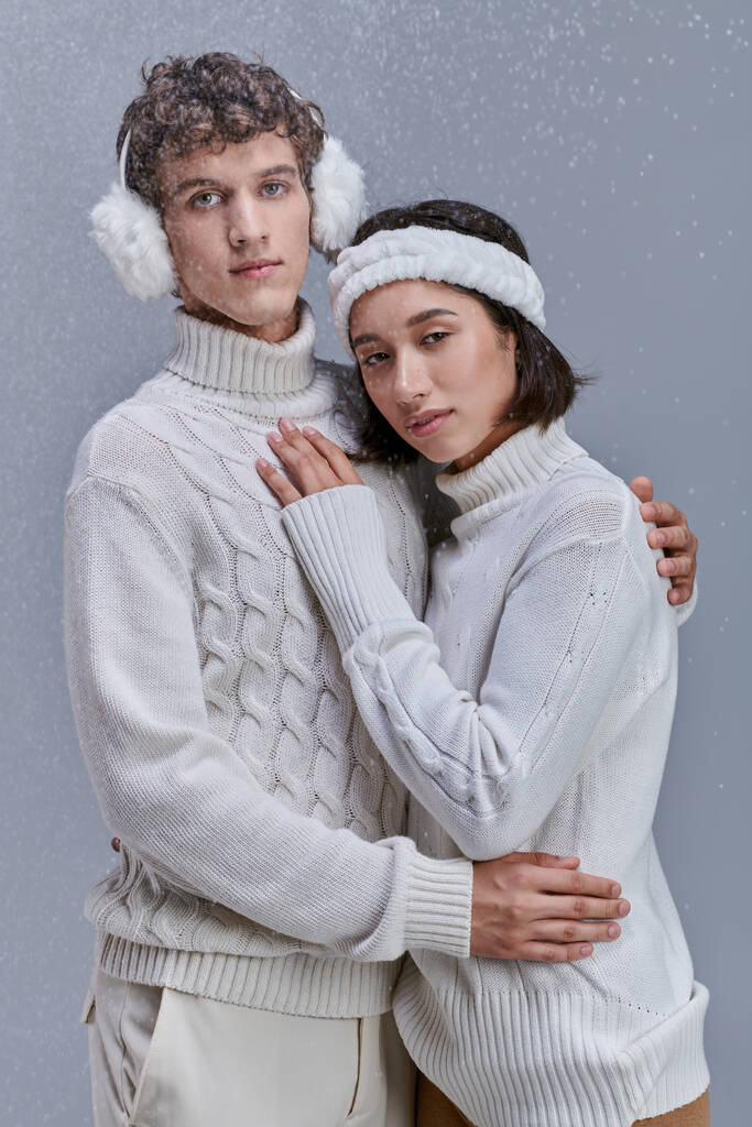 pareja interracial en suéteres blancos abrazando bajo la nieve cayendo sobre fondo gris, moda de invierno - Foto, Imagen